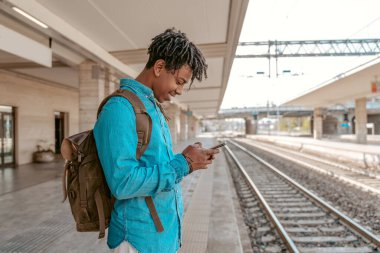 Message. African guy with backpack standing sideways to camera near railway looking joyfully at smartphone screen