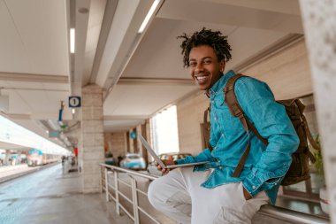 Good day. Smiling african american guy with backpack on back sitting with laptop near railroad looking at camera