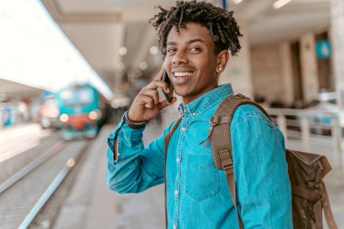 Waiting for train. Young dark-skinned man with backpack talking on smartphone standing near railroad smiling at camera