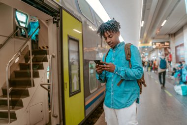 Information. African guy with backpack looking attentively at screen of smartphone standing near open entrance to double-decker train