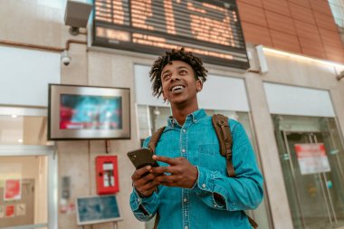 Important information. Dark-skinned smiling guy with smartphone and backpack looking up standing indoors in station with back to timetable