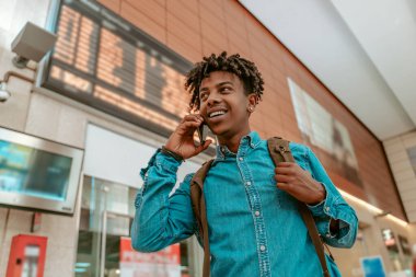 Phone call. Dark-skinned stylish guy with backpack talking on smartphone at station standing with his back to timetable