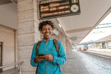 Travel. Dark-skinned cheerful guy with smartphone smiling at camera standing outdoors near railway on fine day