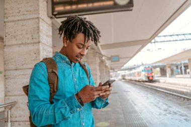 Useful information. Dark-skinned stylish guy with backpack standing on railway platform looking into smartphone on sunny day