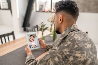 Man in the camouflage uniform sitting at the kitchen table and holding the photo in his hands