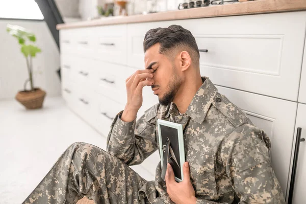 Unhappy military man sitting on the kitchen floor and pressing the picture against his chest