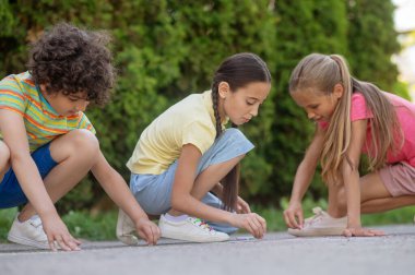 Leisure, drawing. Two long-haired girls and curly-haired boy in bright casual clothes are drawing on asphalt in fresh air