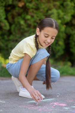 Lucky drawing. Dark-haired smiling girl with pigtails in tshirt and breeches carefully examining drawing on pavement