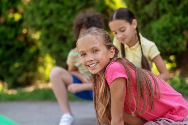 Great day. Joyful long-haired girl in pink tshirt with friends in green park spending leisure time on fine day