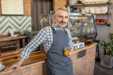 Mid aged man in apron standing near cafe counter with a glass of juice in hand