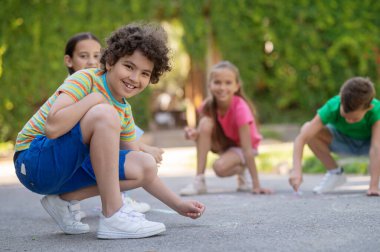 Favourite hobby. Curly smiling boy in tshirt and shorts drawing with friends with crayons in park on summer day