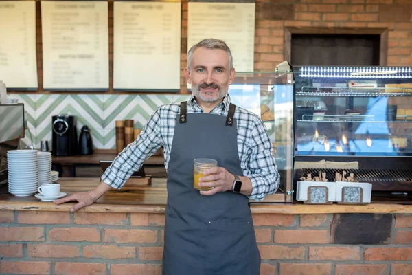 Mid aged man in apron standing near cafe counter with a glass of juice in hand