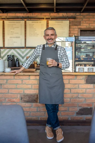 Mid aged man in apron standing near cafe counter with a glass of juice in hand