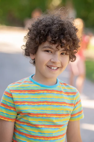 Happy time. Friendly cute smiling boy with dark curly hair in striped tshirt standing in park on fine day