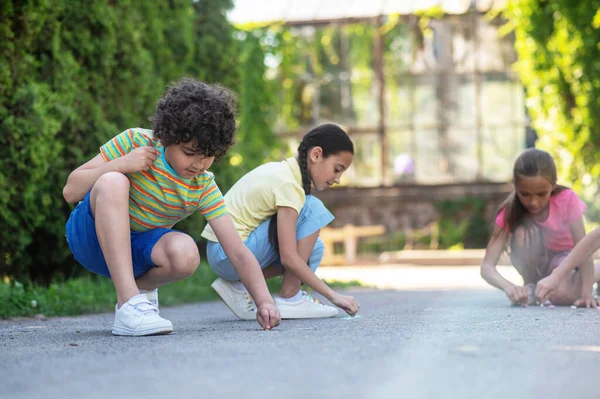 Drawing on asphalt. Focused young friends drawing with crayons on road in green park on sunny day