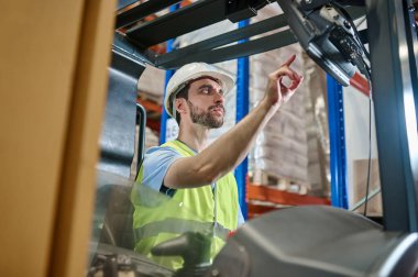 Serious focused young warehouse employee in a reflective vest and helmet starting the lift truck