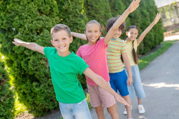 Happy childhood. Cheerful smiling boys and girls with arms outstretched to sides playing in park on summer day