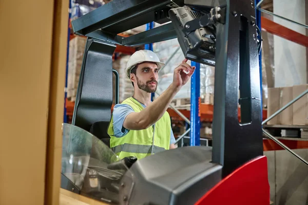 Serious concentrated young storehouse worker in a helmet and reflective vest starting an electric forklift