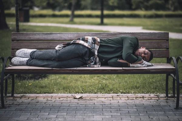 Social problems. African american young man lying sleeping on bench in city park during day