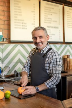 Smiling cheerful coffee shop worker dressed in the apron cutting an orange on the countertop