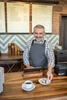 Waist-up portrait of a smiling friendly cafe worker putting a cup of espresso and chocolate crinkle cookies on the counter