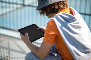 Back view of a male person in the hat with the tablet computer in his hands sitting outside