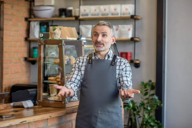 Friendly mature gentleman dressed in the apron standing in the coffee shop and greeting his clients