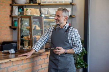Smiling happy cafe worker with the smartphone in his hands leaning on the serving counter