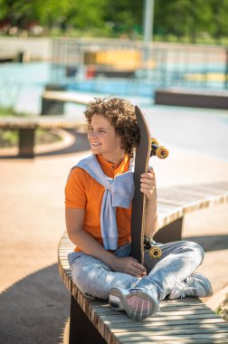 Smiling boy sitting cross-legged on the bench with his skateboard and looking into the distance