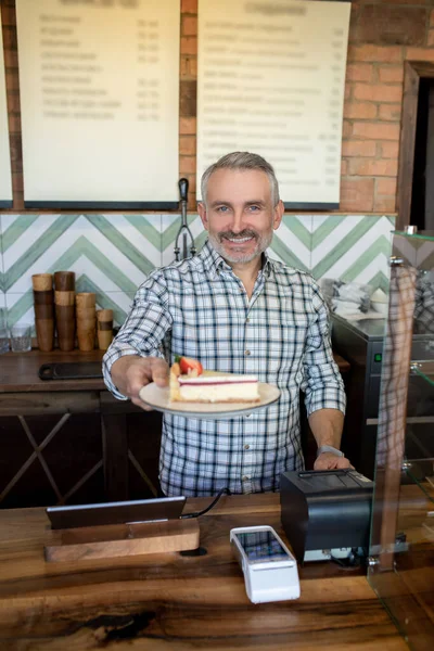 Waist-up portrait of a smiling pastry shop employee handing a piece of cake to his customer