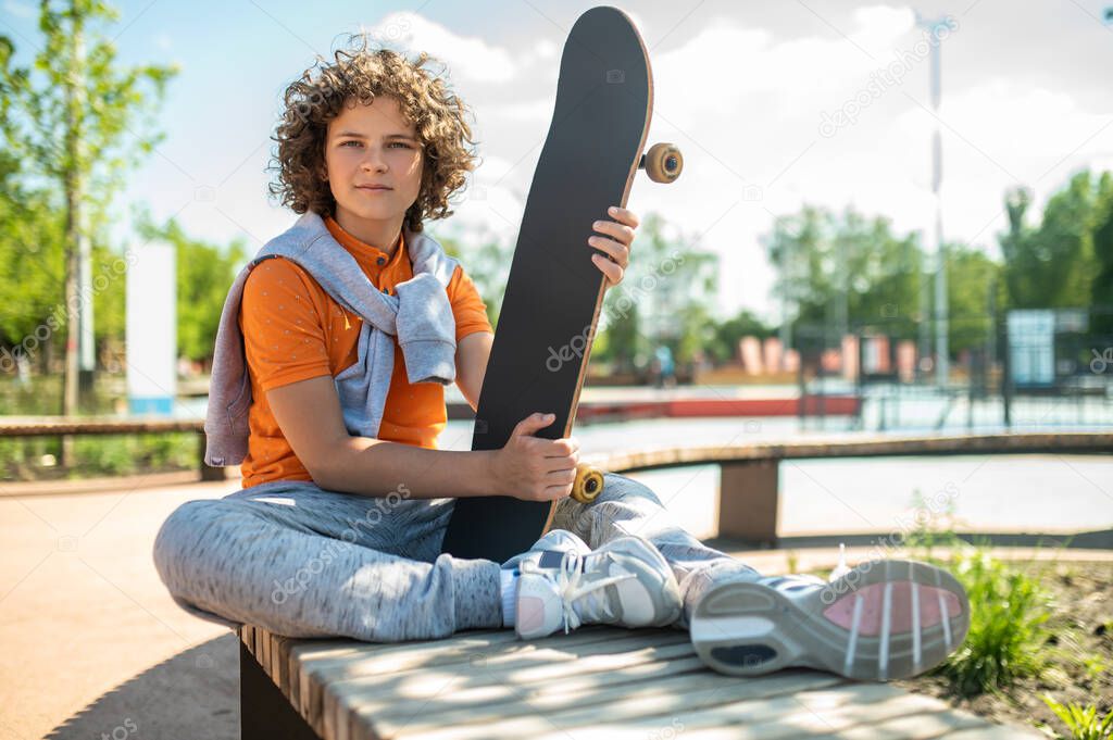 Tranquil cute adolescent skateboarder sitting on the bench and holding his board in the hands