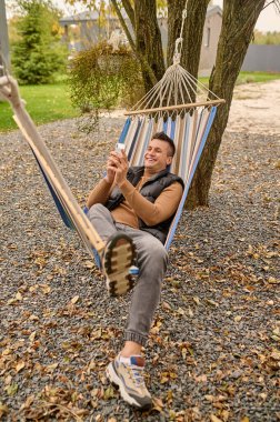 Smiling pleased guy lying in the hammock and looking at the smartphone in his hands