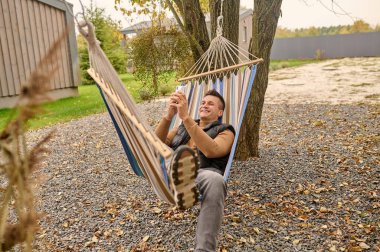 Smiling happy handsome male person lying in the hammock with the cellphone in his hands