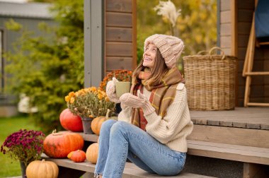 Joyous woman seated on the wooden porch steps with a mug in the hands looking up at something