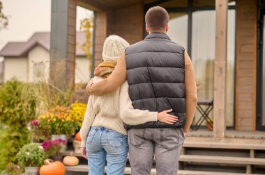 Back view of a woman and a man in warm clothes standing before a wooden cottage