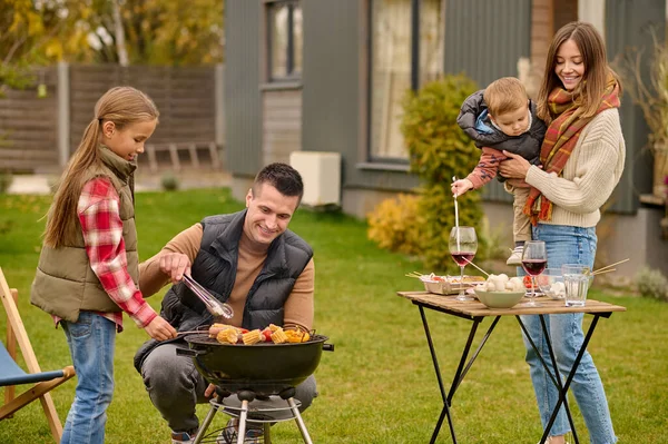 Teenage girl and her dad cooking food on the grill while her mother playing with a younger child
