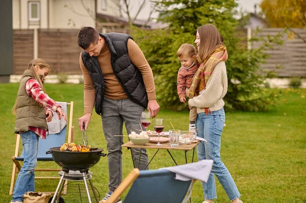Adult male grilling veggies on the grill in the presence of his spouse and children
