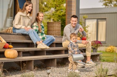 Sulama kabı olan küçük bir çocuk verandada oturan kız kardeşi ve neşeli ebeveynleriyle kameraya poz veriyor.