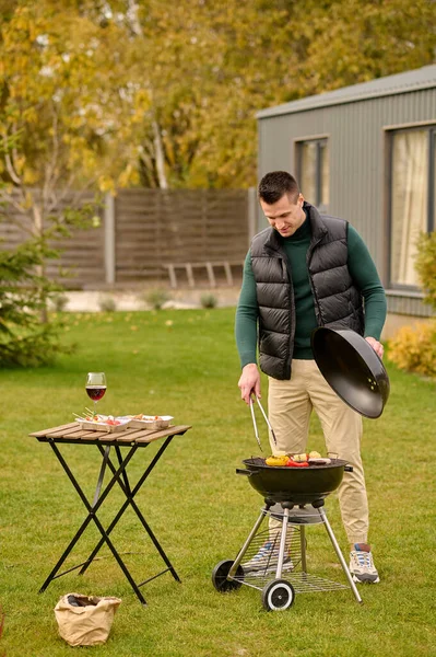 Full-size portrait of a guy standing before the open brazier in the backyard while grilling veggies and sausage