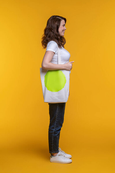 Casual style. Smiling long-haired woman in jeans with bag on speck standing sideways to camera on yellow background