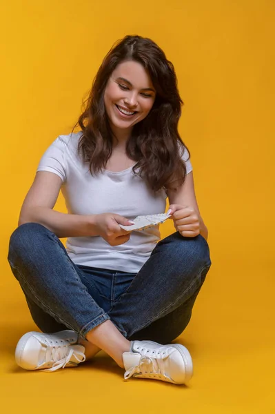 Good moments. Smiling young woman in jeans looking at letter in hands sitting cross-legged on floor on yellow background