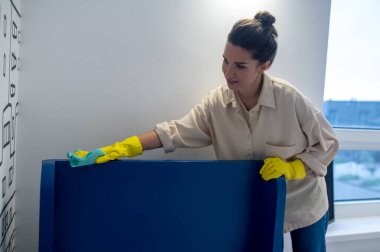 Koyu renk saçlı güzel bir kadın. Yatak başlığının üst korkuluğunu mikrofiber bezle silmeye odaklanmış.