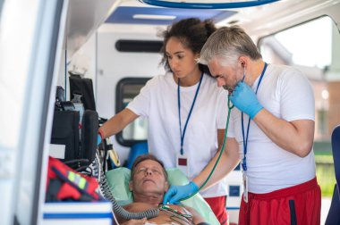 Kadın meslektaşı EKG monitörüne bakarken odaklanmış sağlık görevlisi erkek hastanın kalp atışlarını dinliyor.