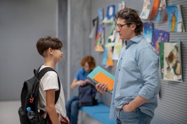 Sınıf arkadaşı koridorda bankta otururken sırt çantalı bir öğrenci bir öğretmenle iletişim kuruyor.