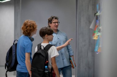 Sırt çantalı okul çocukları koridor duvarında durup öğretmenlerinin açıklamalarını dinliyorlar.