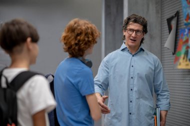 Elinde bir yığın ders kitabı olan bir öğretmen koridorda dikilip okul çocuklarıyla konuşuyor.