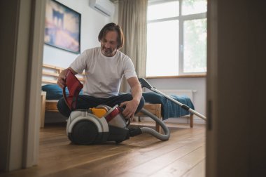 Ev işi. Adam yerde oturuyor ve elektrik süpürgesini topluyor.
