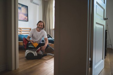 Ev işi. Adam yerde oturuyor ve elektrik süpürgesini topluyor.