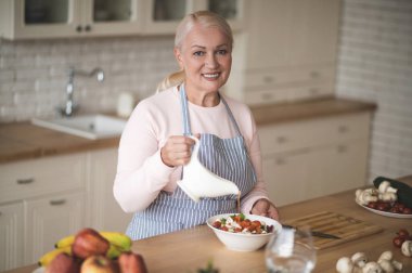 Önlükteki gülümseyen mutlu kadın aşçı. Kasedeki doğranmış sebzelere sos ekliyor.