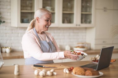 Önlükteki mutlu, olgun yemek vlogger 'ı dizüstü bilgisayar ekranından önce kasede sebze salatası tutuyor.